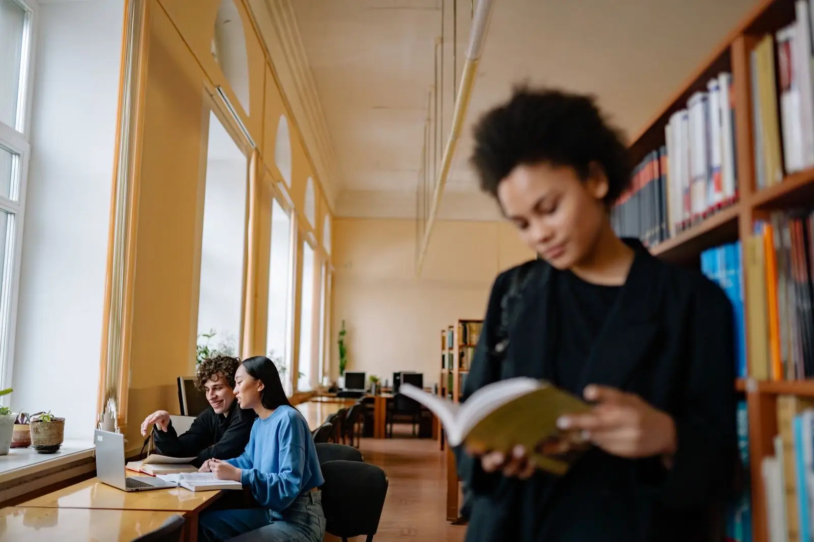 A doctoral student reading in a university library while others work on laptops nearby